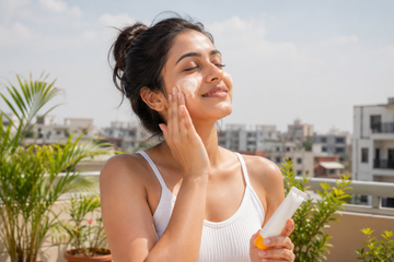 Young Indian woman applying sunscreen on her face outdoors under bright daylight on a sunny rooftop, demonstrating daily sun protection for Indian skin and climate.